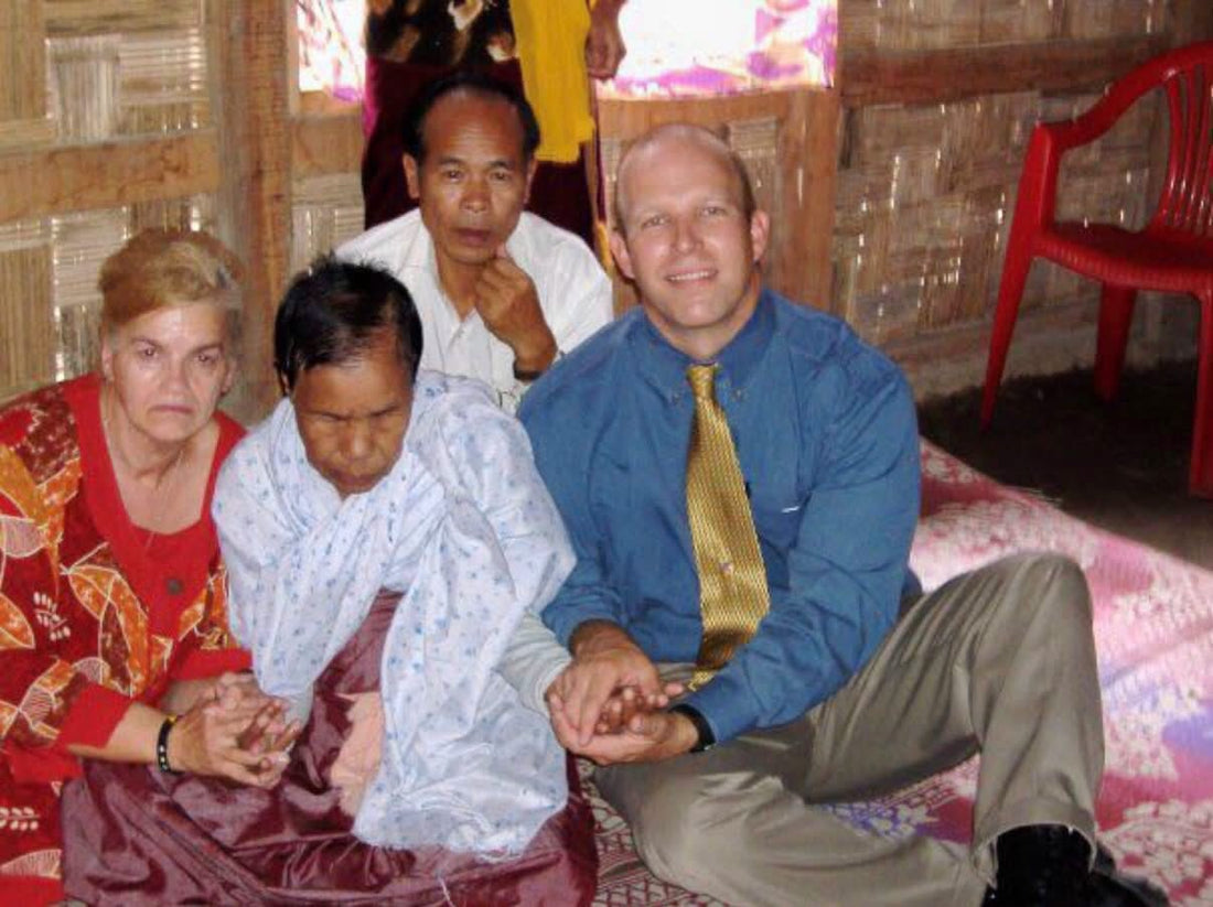A woman seated between two visitors holding her hands inside a home, with another man seated behind them.